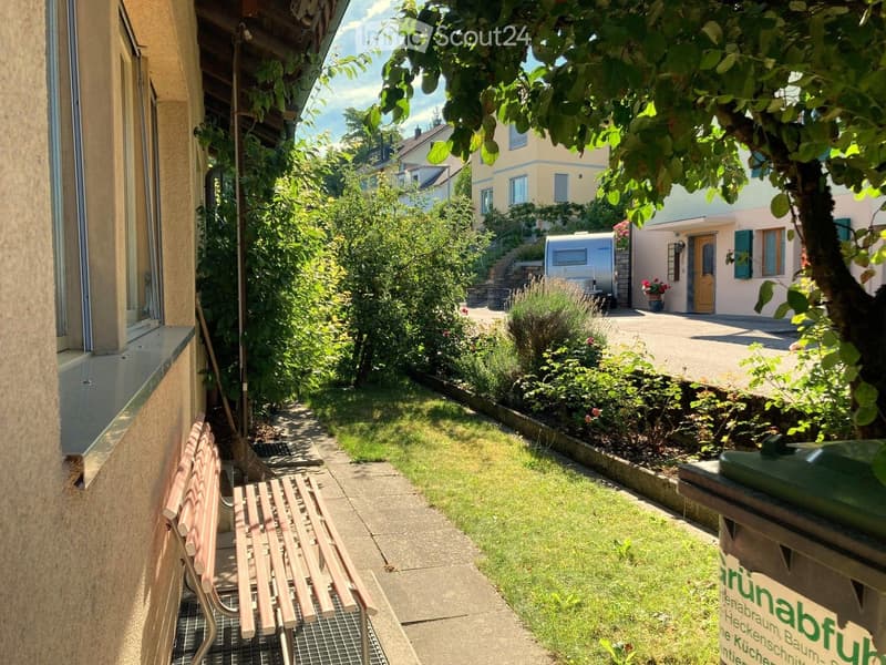A garden area with a bench, a tree, and a trash bin. The garden is surrounded by a concrete pathway and has a grassy lawn. There are plants and flowers in the garden, and a building with a door and a window is on the right side. A camper van is parked on