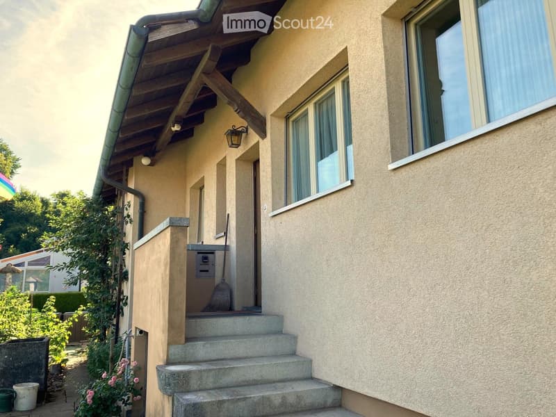A beige house with a wooden roof, concrete steps, and a lamp. A broom is leaning against the wall next to the door. A bucket and a plant are on the left side.
