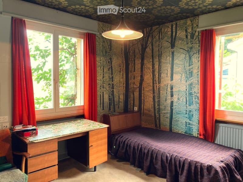 Small room with a desk, bed, and a wallpapered wall. The room has two windows with red curtains.