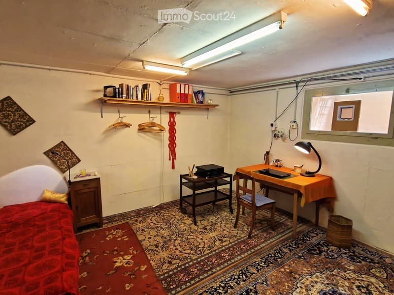 Small furnished room with a bed, desk, chair, and shelves. Features patterned rugs, a window, and basic lighting.