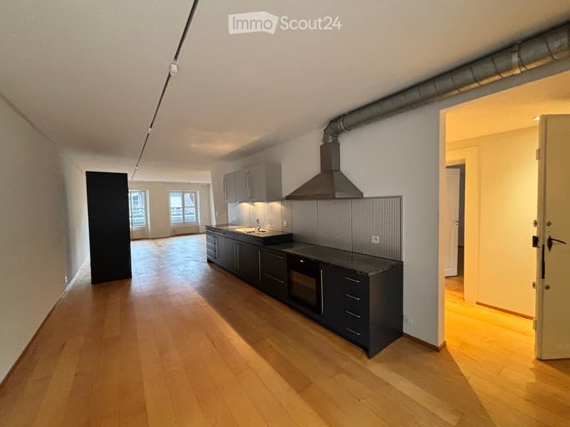 Modern kitchen with dark cabinets, a sink, oven, and range hood. The floor is wooden, and there are windows in the background.
