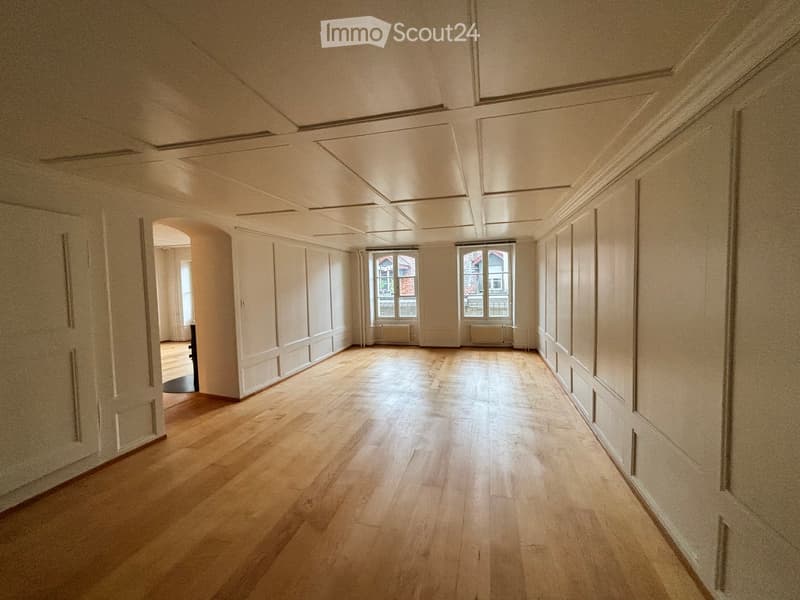 Empty hallway with wooden flooring, white walls, and large windows at the end, allowing natural light to enter.
