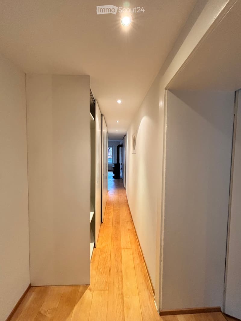 A narrow hallway with light wooden flooring and white walls, featuring recessed ceiling lights and a visible room at the end.