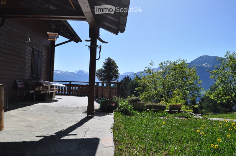 A wooden deck area with a table and chairs, overlooking a scenic mountain view with lush greenery and flowers.