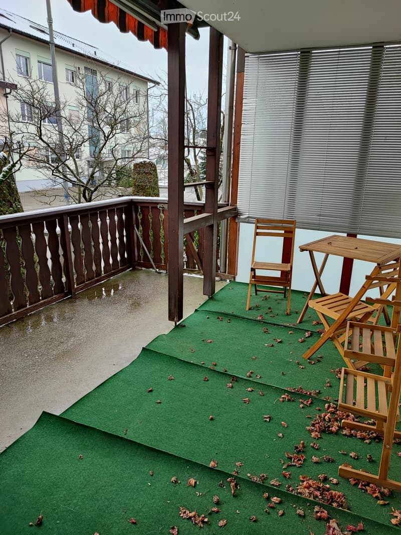 Balcony with green artificial turf, wooden table, and chairs, overlooking a residential area with neighboring buildings.
