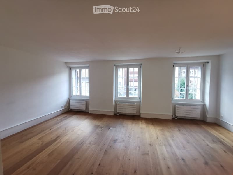 Empty room with wooden floors, white walls, three windows, and white radiators