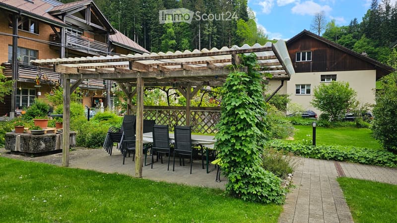 A well-maintained garden with a wooden pavilion, a table and chairs, flower pots, and lush greenery.