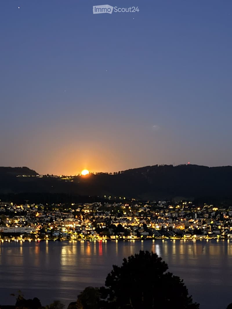 Nighttime view of a cityscape with lights reflecting on water, distant mountains, and a setting sun.