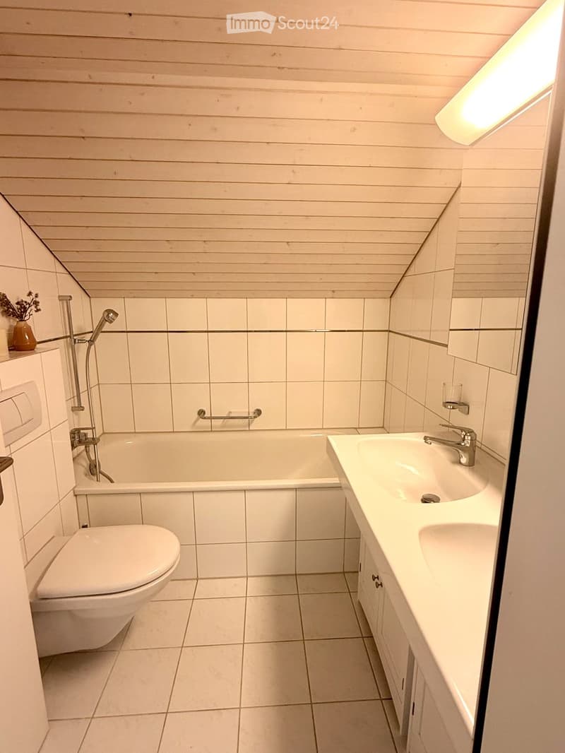 Bright bathroom with white tiled walls and floor, featuring a bathtub, toilet, sink, and showerhead. A small vase with flowers sits on a shelf next to the toilet.