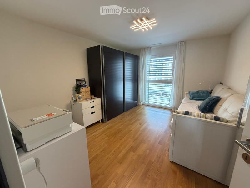 Cozy bedroom with wooden flooring, a single bed with pillows, a large wardrobe, a nightstand with a picture frame and plant, and a printer on a small table.