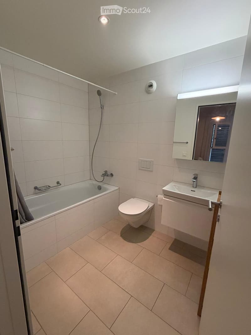 Modern bathroom with a bathtub, toilet, sink, mirror, and shower. Light-colored tiles and warm lighting.