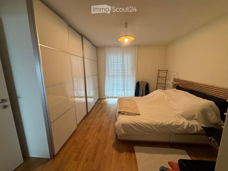 A modern bedroom with a large bed, a wardrobe, a chair, and a window with curtains. The room features wooden flooring and a hanging lamp.