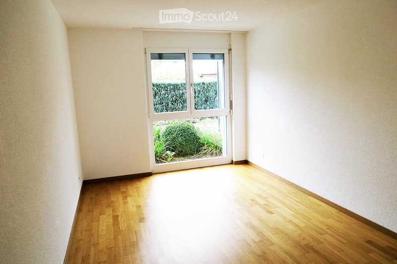 Empty room with wooden flooring and white walls, featuring a large window with a view of greenery outside.