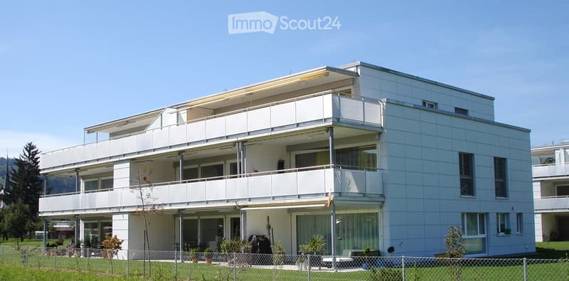 Modern white apartment building with multiple balconies and large windows, surrounded by a small garden area with potted plants.