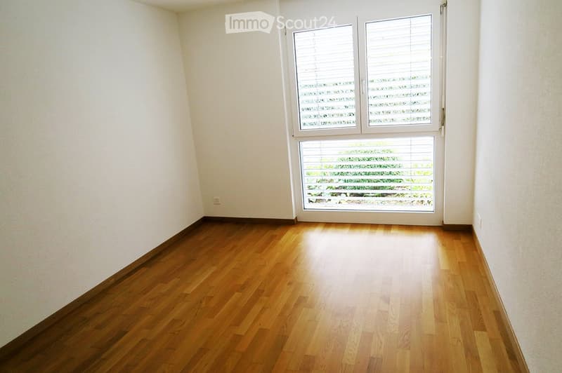 Empty room with wooden flooring and white walls, featuring a large window with blinds.