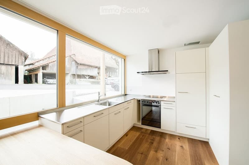 Modern kitchen with white cabinets, stainless steel appliances, wooden flooring, and large windows overlooking a street.