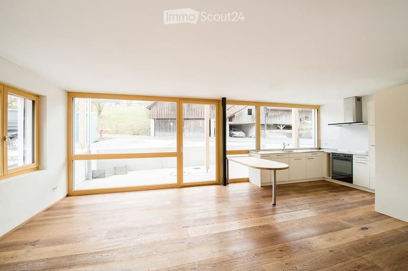 Bright, modern kitchen with light wooden flooring, large windows, and white cabinetry. Features a central island table, sink, and oven.
