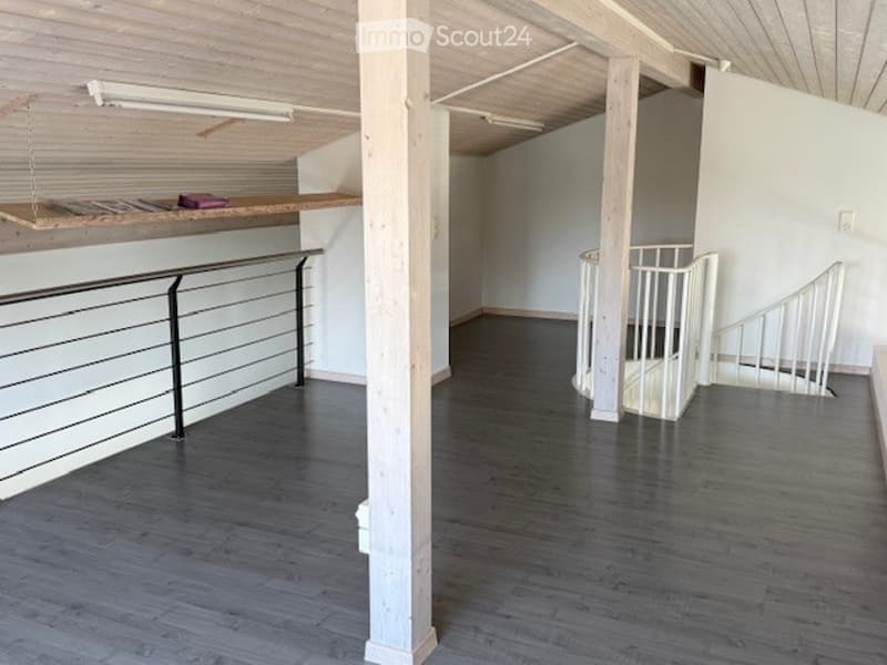 Open loft-style space with exposed wooden beams, dark grey flooring, white walls, and a staircase leading to an upper level.