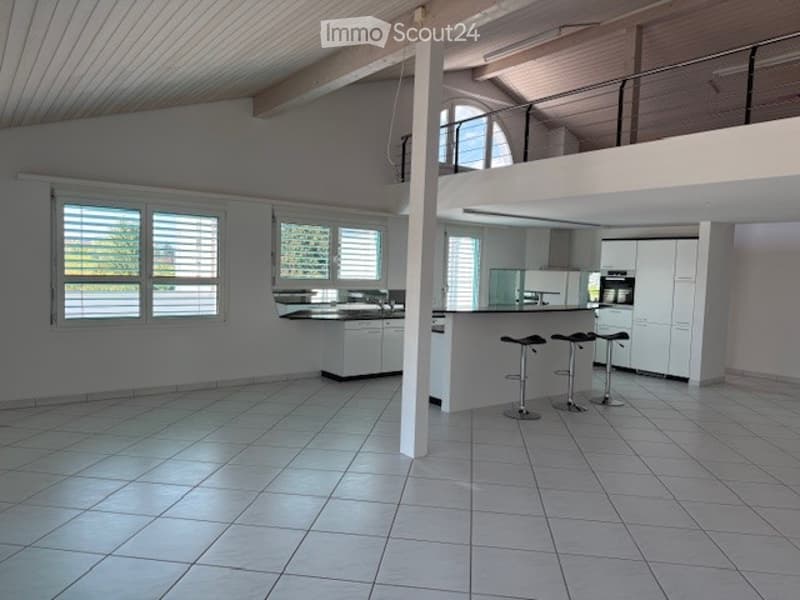 Open-plan kitchen and living area with white tiled flooring, white kitchen cabinets, a central island with bar stools, and large windows with blinds.