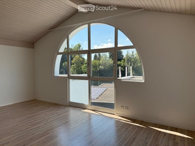 Bright, empty room with wooden flooring and large arched window offering a view of the outdoors.