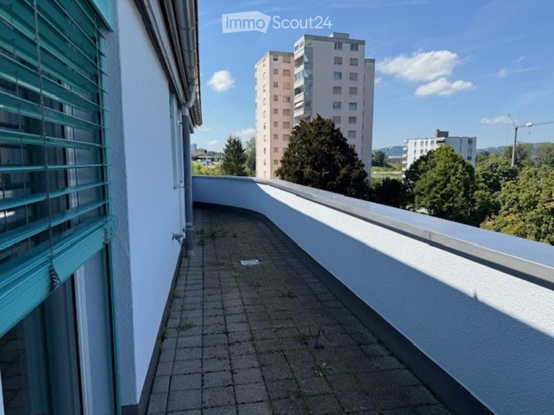 Balcony with tiled floor, white walls, and a view of surrounding buildings and greenery.