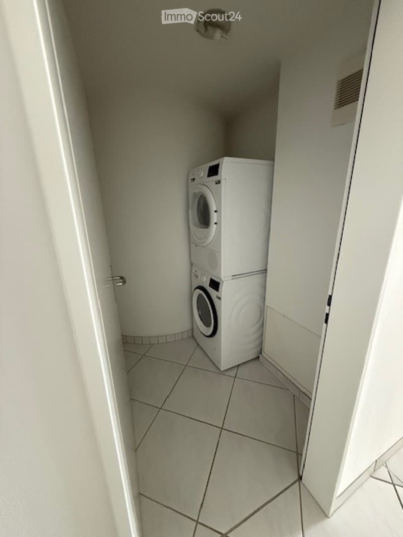 Small, tiled room with a white washing machine and dryer stacked vertically against the wall.