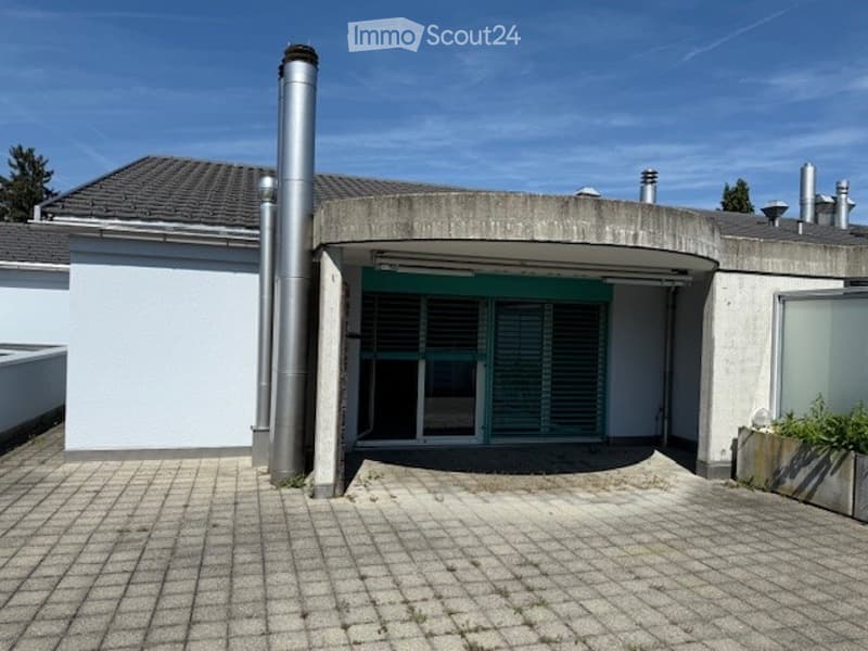 Modern single-story building with a flat roof, white walls, and a central entrance with green shutters. The entrance features a covered area with a concrete floor and a large window. The building has multiple ventilation pipes on the roof.
