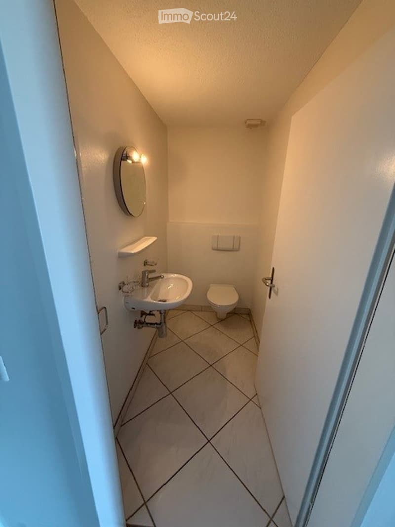 Small, modern bathroom with white tiled floor and walls, a sink, toilet, mirror, and a small shelf.