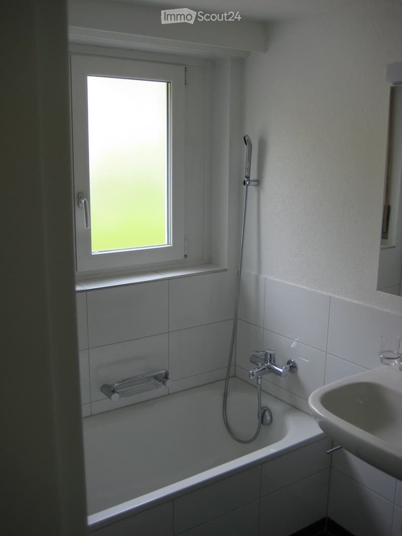 Modern bathroom with white tiled walls, bathtub, shower hose, sink, and a window with frosted glass.