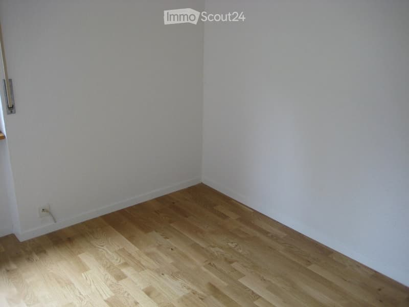 Empty room with light wooden flooring and white walls, featuring a visible electrical outlet and a small metal fixture on the wall.