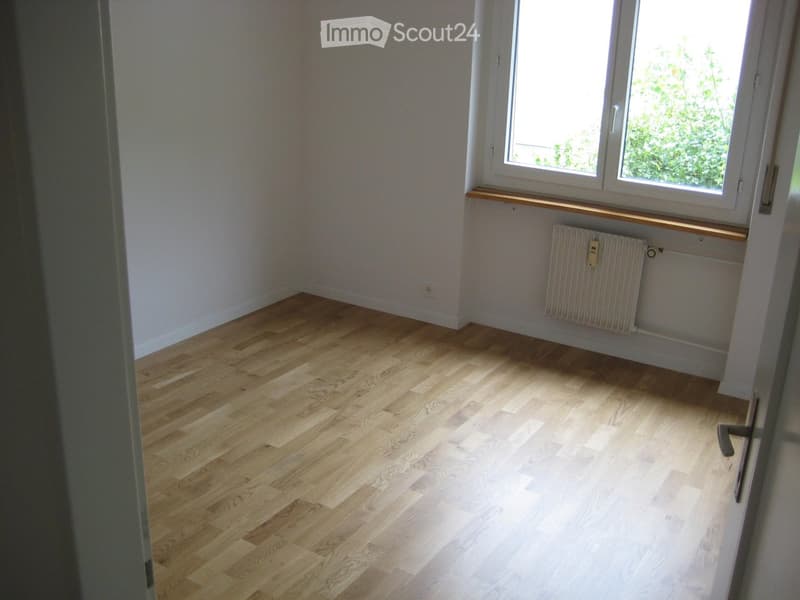 Empty room with light wooden flooring, white walls, a window with a radiator below, and a door frame visible on the left side.