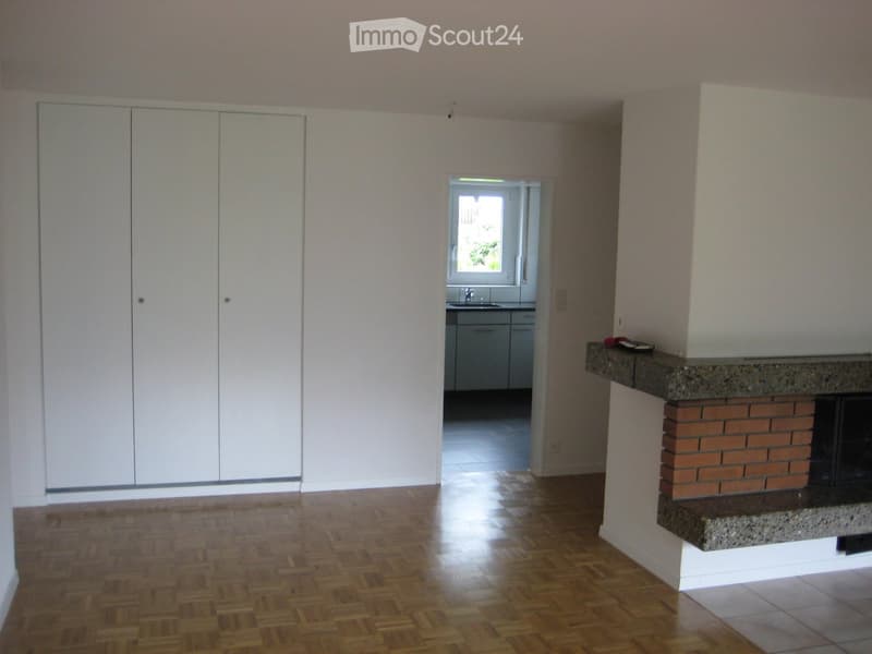 Empty room with wooden flooring, white walls, and a fireplace with a stone surround. A doorway leads to a kitchen area with cabinets and a sink.