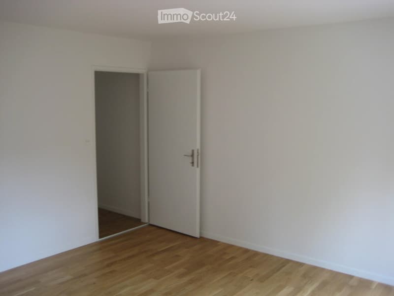 Empty room with light-colored walls and wooden flooring, featuring an open door leading to another space.