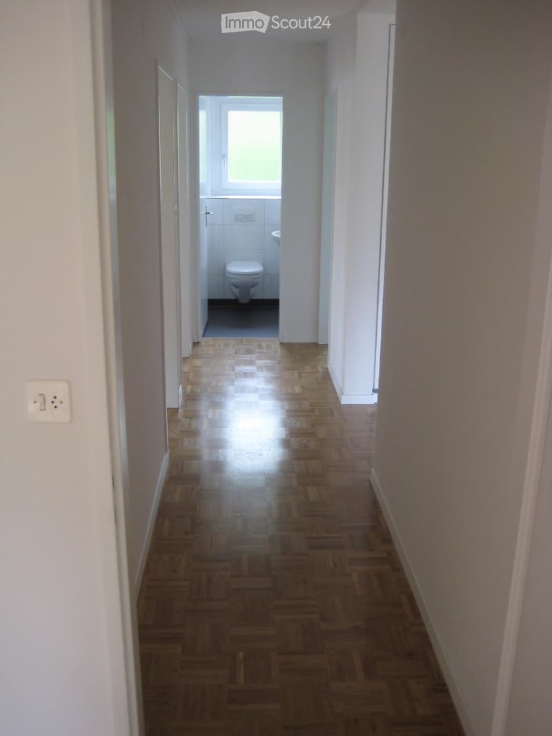 A narrow hallway with wooden flooring and white walls, leading to a small bathroom with a toilet and a window.