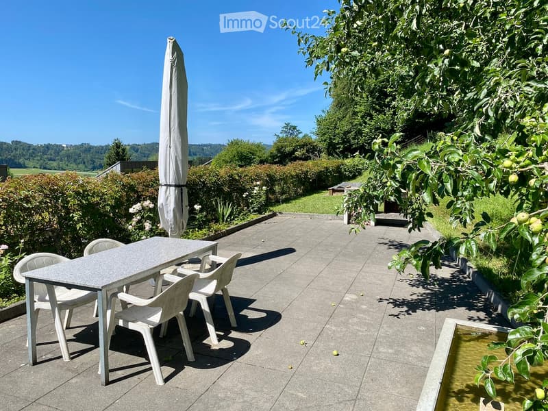 Outdoor patio with a grey table, white plastic chairs, and a closed umbrella. The area is surrounded by greenery, including a tree with fruit and a small water feature.