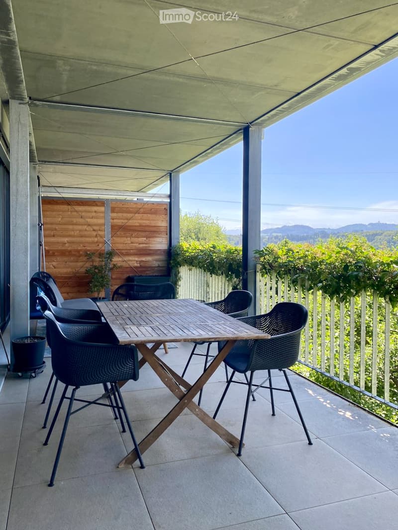Balcony with a wooden table and black chairs, covered by a canopy, overlooking a scenic view of greenery and mountains.