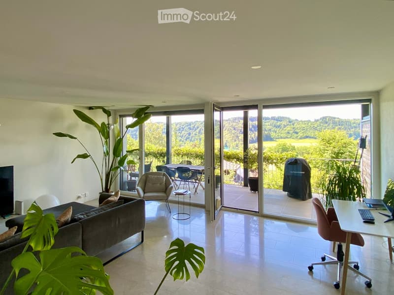 Modern living room with large sliding glass doors opening to a balcony, offering a scenic view of greenery. The room features a grey sofa, a beige armchair, a small side table, a desk with a chair, and several potted plants.