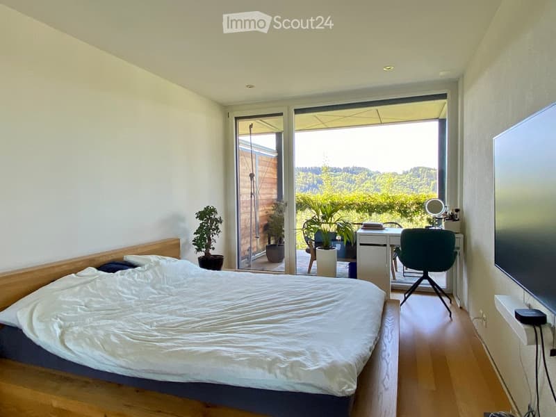 Bright bedroom with a wooden bed, desk, chair, and large sliding glass doors opening to a balcony with a scenic view.