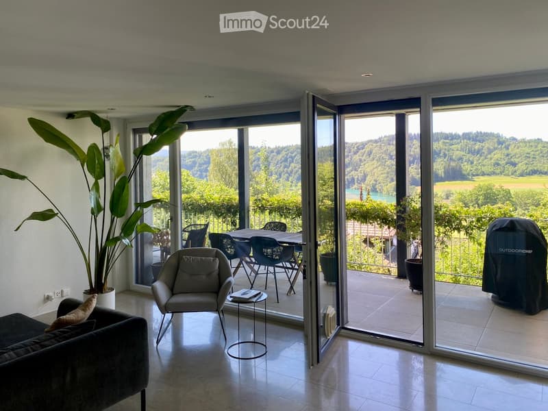 Modern living room with large sliding glass doors opening to a balcony, featuring a sofa, armchair, small table, and potted plant. The balcony includes a dining table and chairs with a scenic view of a valley and lake.