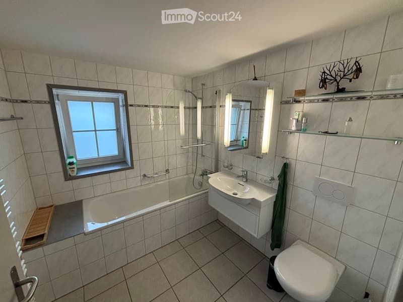 Bathroom with tiled walls and floor, featuring a bathtub, sink, toilet, mirror, shelves, and a window with a small wooden step stool.