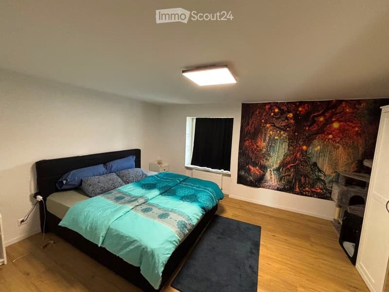 Cozy bedroom with a large bed, blue and grey pillows, teal blanket, nightstand, mirror, and a large wall tapestry.