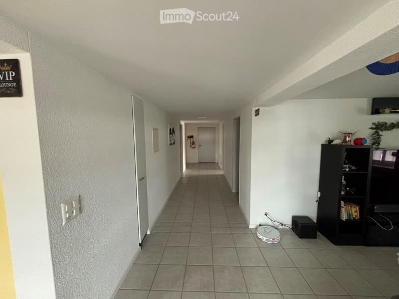 A long, narrow hallway with tiled flooring, white walls, and several doors leading to different rooms. A small shelf with books and a speaker is visible on the right side.