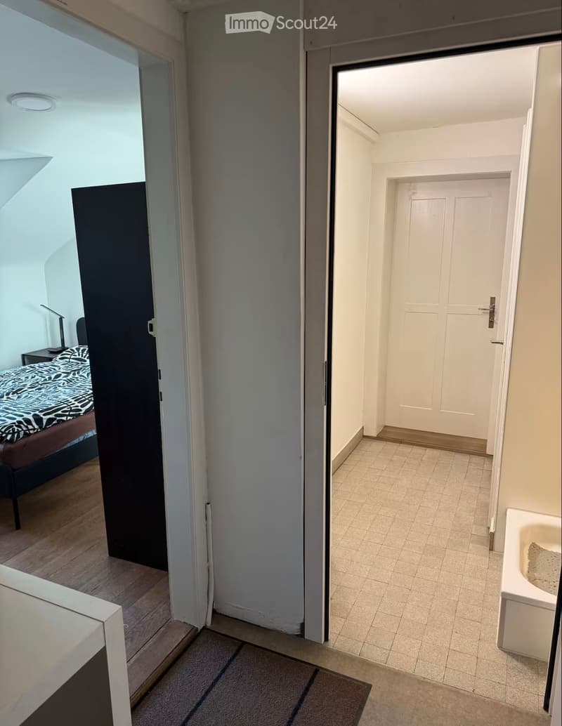 Hallway leading to a bedroom and a bathroom. The bedroom has a bed with a patterned duvet and a nightstand. The bathroom has a bathtub and tiled flooring.