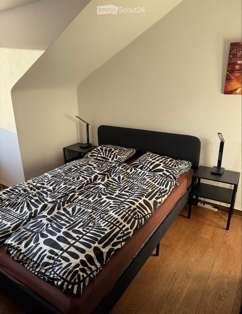 Bedroom with a single bed, black and white patterned duvet, two nightstands with lamps, and wooden flooring.