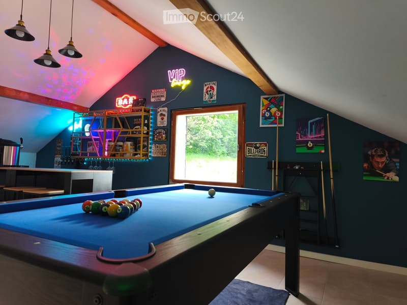 A well-lit room with a pool table, a bar area, shelves with bottles and glasses, neon signs, and artwork on the walls.