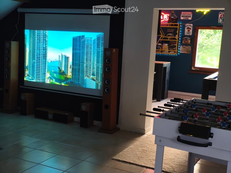 A modern entertainment room with a large screen displaying a cityscape, multiple speakers, a football table, and a small table with a window view.