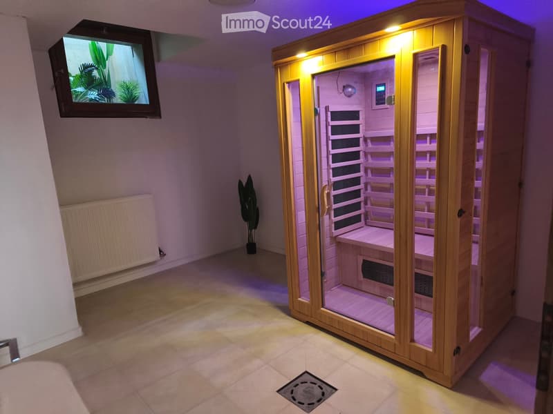 A modern sauna room with wooden sauna benches, a control panel, a TV screen, a radiator, and a potted plant.