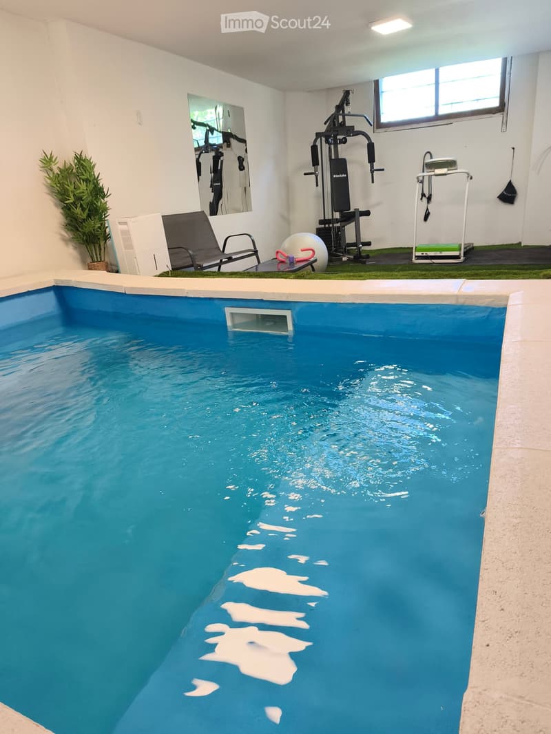Indoor swimming pool with clear blue water, surrounded by exercise equipment, a lounge chair, and a small potted plant.
