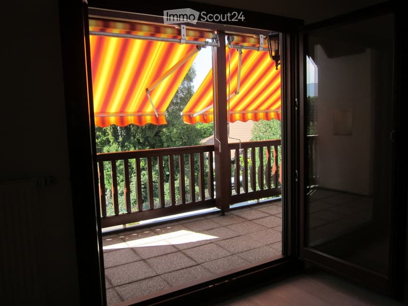 A balcony with an awning, wooden railing, tile floor, and an open glass door leading to the inside of a building. Outside the balcony, there are trees and a house.