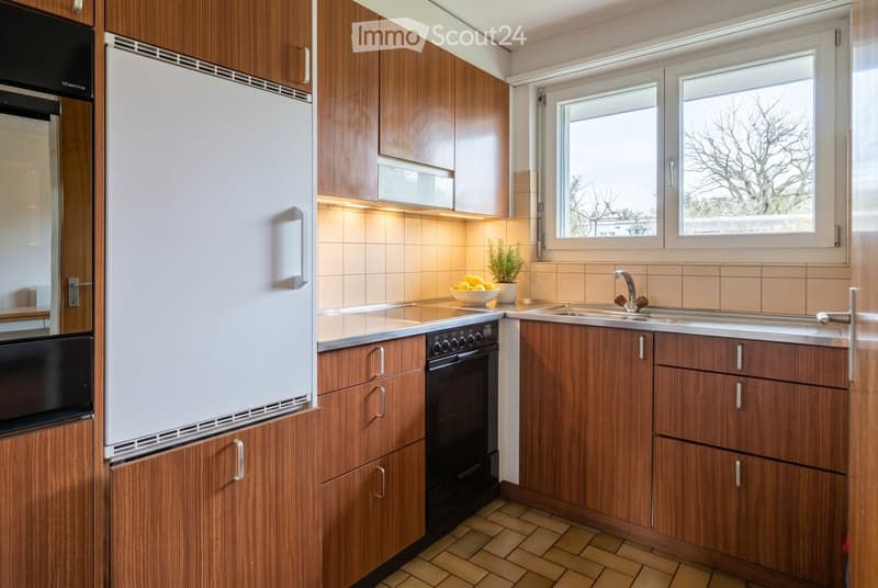 Modern kitchen with wooden cabinets, stainless steel appliances, and a tiled backsplash. Features a refrigerator, oven, sink, and a small potted plant on the counter.